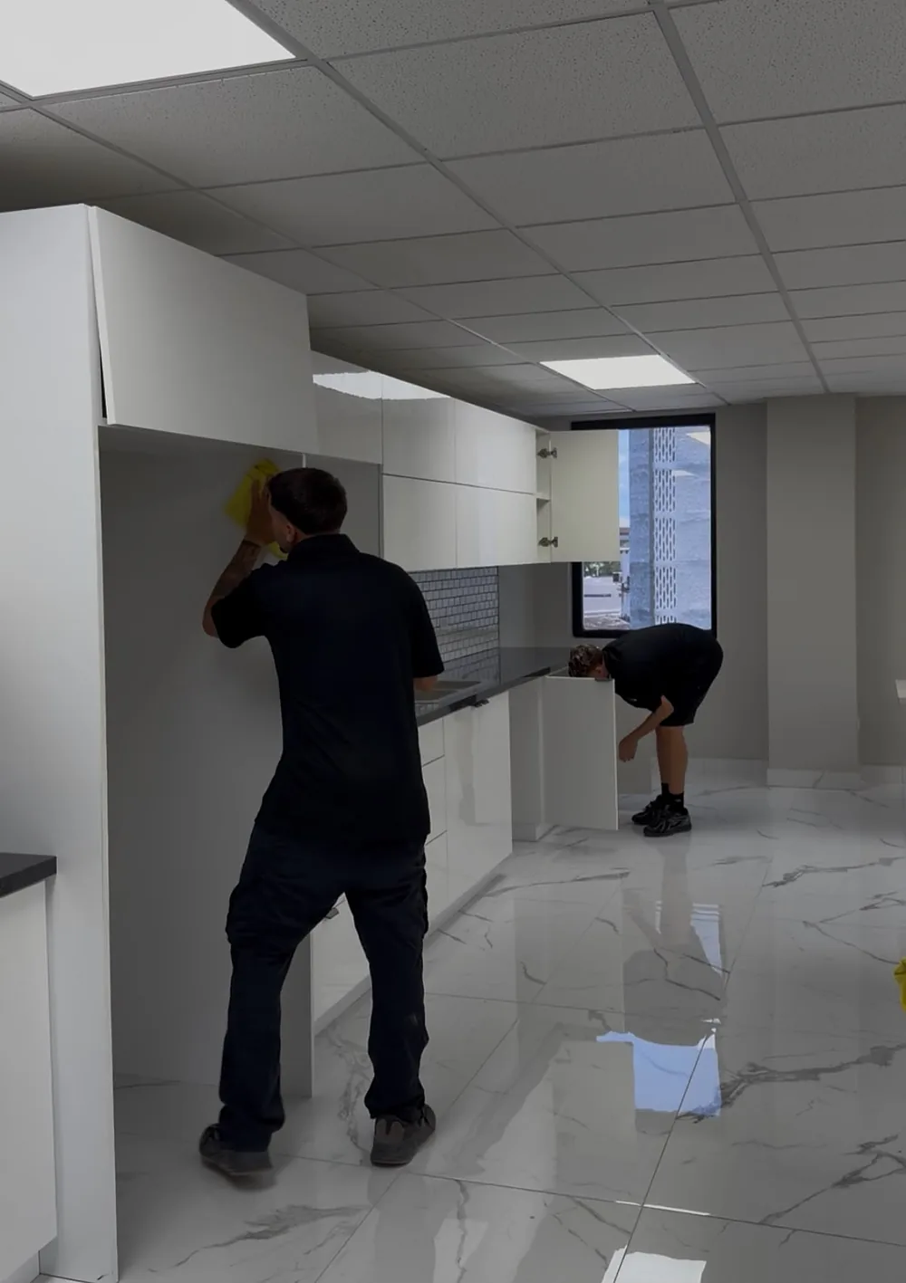 Dos empleados de Clean Vibes limpiando la cocineta de una oficina, con gabinetes blancos y piso de mármol